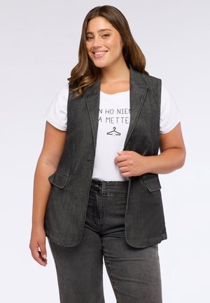 Lachende vrouw met lang bruin haar draagt een mouwloze donkergrijze blazer over een wit shirt met bedrukt tekst en donkergrijze jeans.