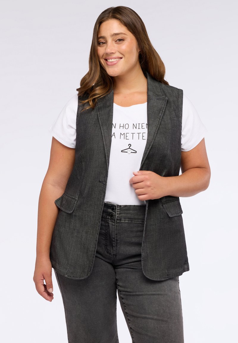 Donna sorridente con lunghi capelli castani indossa un blazer senza maniche di un grigio scuro sopra una camicia bianca con testo stampato e jeans grigi scuri.