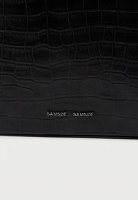 Μαύρο δέρμα με σχέδιο κροκοδείλου και λαμπερό λογότυπο "SAMSØE SAMSØE". Ομαλή υφή με διακριτές λέπιες και λεπτομέρειες ραψίματος.