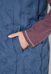 Hand in der Tasche einer blauen gesteppte Jacke, die über einem langärmeligen bordeauxroten Hemd mit sichtbaren Nähten an den Ärmelbündchen getragen wird.