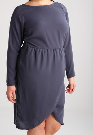 Laivastonsininen polvipituinen mekko, jossa on pitkät hihat, kerrostettu vyötärö ja päällekkäinen etuhalkio, pukeutuneena henkilöön, jonka kädet ovat rentoina.