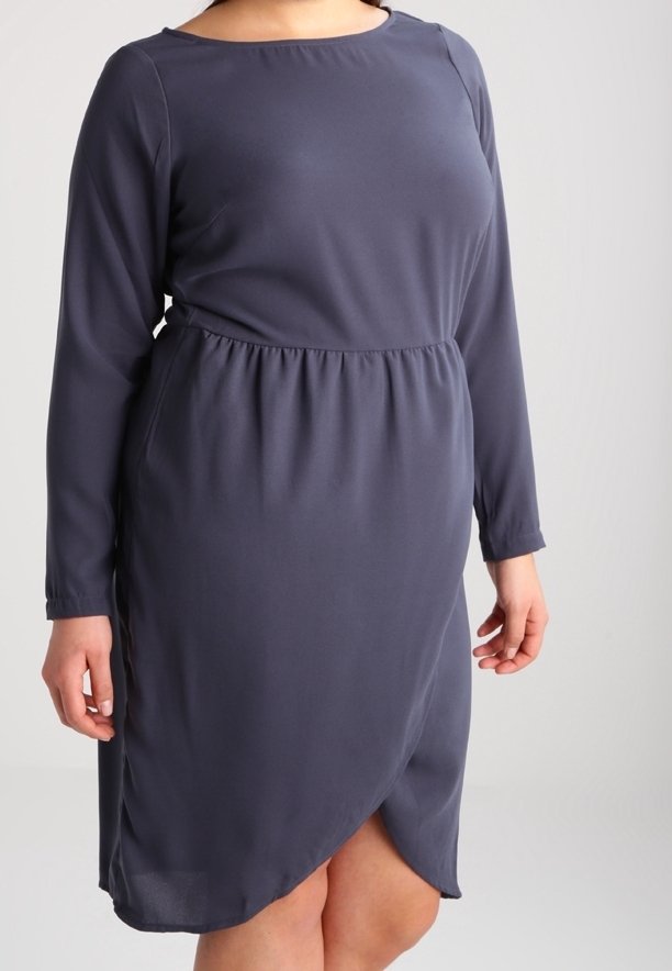 Laivastonsininen polvipituinen mekko, jossa on pitkät hihat, kerrostettu vyötärö ja päällekkäinen etuhalkio, pukeutuneena henkilöön, jonka kädet ovat rentoina.