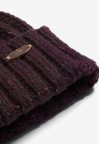 Lila kötött sapka bordás textúrával és fémből készült címkével, fehér szálak pöttyözésével kiemelve.