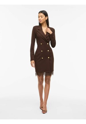 Femme portant une robe blazer marron cintrée à double boutonnage avec des boutons dorés et une bordure en dentelle, debout et regardant sur le côté.
