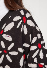 Black knit sweater featuring large white daisies and red centers, with a small branded logo tag stitched on the lower left side.