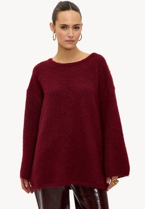 Femme aux cheveux foncés attachés en chignon, portant des boucles d'oreilles anneau dorées, un pull bordeaux ample et duveteux, ainsi qu'un pantalon noir brillant, debout devant un fond blanc.