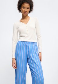 Γυναίκα που φοράει κρεμ μακρυμάνικη μπλούζα henley και μπλε παντελόνι υψηλής μέσης με ροζ κάθετες ρίγες και λεπτομέρειες με κουμπιά.