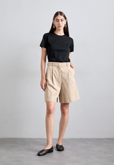 Woman standing against gray wall wearing black short-sleeve shirt, beige knee-length shorts, black woven flats, and statement earrings.