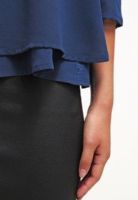 Marinblus med en lagerkant och subtil logotypdetalj, i kombination med en svart, figurnära kjol i ett elegant material.
