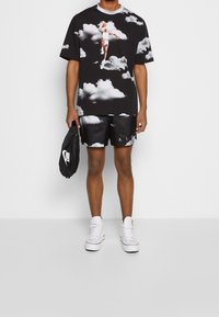 T-shirt noir avec motif nuage et graphique de basket-ball, associé à un short noir avec le texte "AIR", accessoirisé avec un sac noir et des baskets blanches.