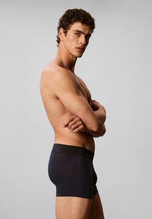 Oberkörperfreier Mann mit lockigem Haar steht im Profil mit verschränkten Armen und trägt dunkle, eng anliegende Boxershorts vor einem einfarbigen hellen Hintergrund.