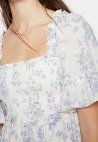 Biała sukienka z lawendowym kwiatowym wzorem, kwadratowym dekoltem, falbaniastrzymy rękawami i dołem, marszczoną talią oraz teksturowanymi akcentami w kropki.