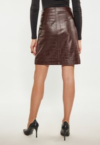 Gonna in pelle marrone con texture a coccodrillo, forma dritta e vita media. Indossata con scarpe con tacco alto nere.