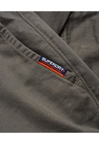 Tessuto grigio con una superficie liscia caratterizzato da un'etichetta blu e arancione "SUPERDRY INTERNATIONAL" cucita in modo prominente sul lato.