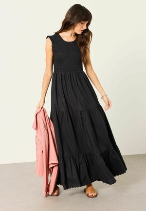 Frau in einem ärmellosen schwarzen gestuften Maxikleid, die eine rosa Jacke hält, braune Sandalen trägt und auf einem neutralen Boden und Hintergrund steht.