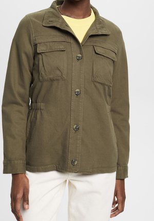 Personne portant une veste boutonnée vert olive avec des poches poitrine et une taille élastique, associée à un pantalon blanc et une chemise jaune en dessous.
