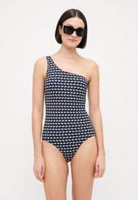 Maillot de bain une épaule en bleu marine avec un motif de nuages blancs. Tissu lisse, coupe ajustée et couverture modérée. Lunettes de soleil noires portées.