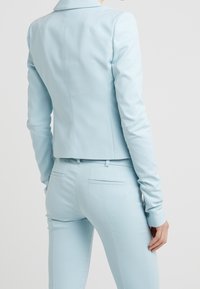 Blazer cintré bleu clair et pantalon assorti, tissu lisse avec un léger brillant, coupe ajustée, doté d'une fente arrière unique et de poignets boutonnés.