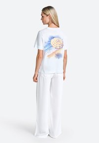 White short-sleeve t-shirt with a colorful watercolor print of a beach umbrella on the back; paired with wide, flowing white pants.