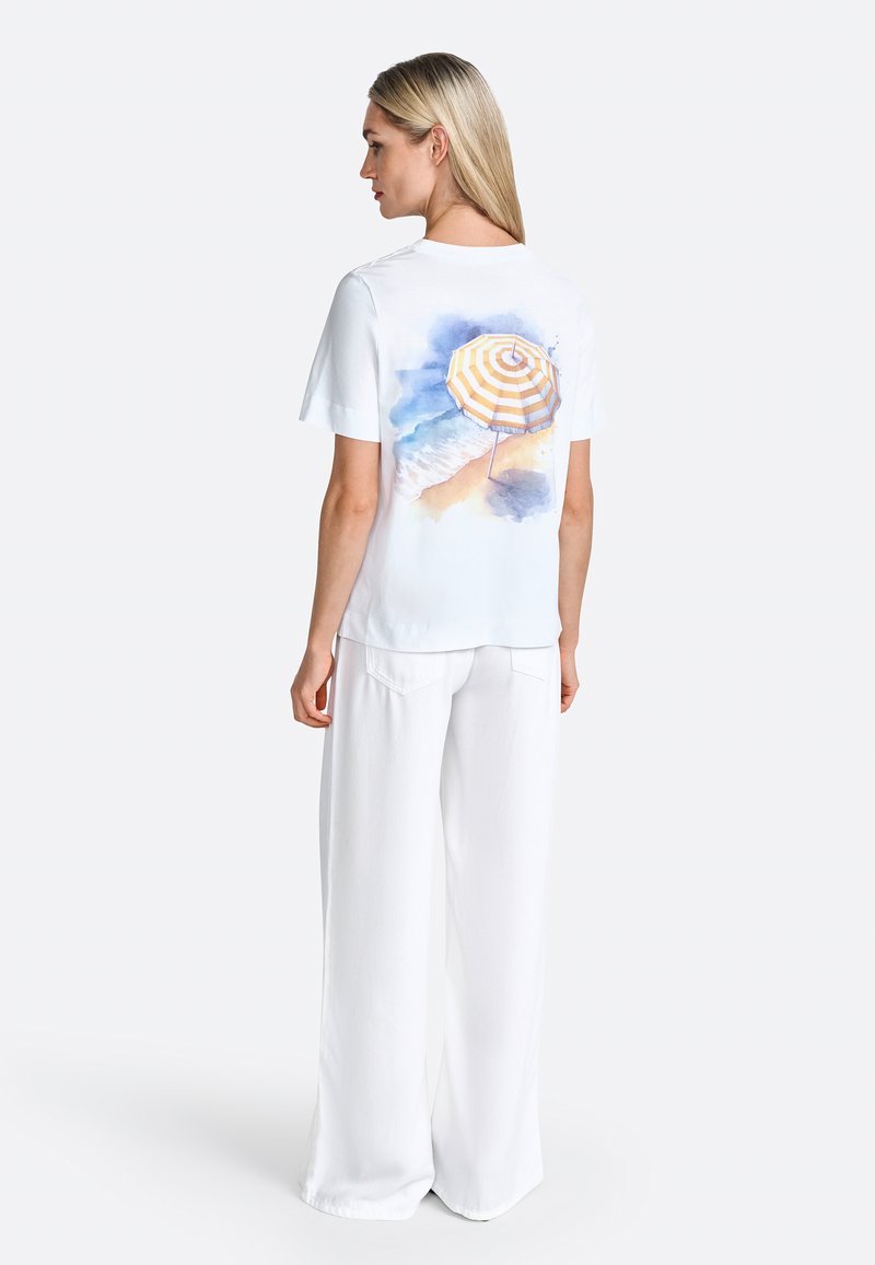 White short-sleeve t-shirt with a colorful watercolor print of a beach umbrella on the back; paired with wide, flowing white pants.
