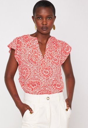 Blusa rossa e bianca con motivo a fantasia, maniche corte a volant, scollatura a V profonda e vestibilità rilassata; indossata con pantaloni bianchi a vita alta.
