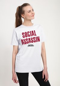 Bílý bavlněný tričko s výrazným červeným textem "SOCIAL ASSASSIN" a menším černým logem níže. Krátké rukávy a volný střih.