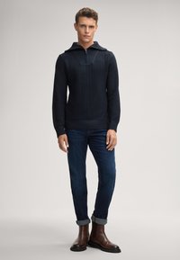 Marinefarbener strukturierter Pullover mit hohem Kragen und Reißverschluss, kombiniert mit dunkelblauen Jeans und braunen Stiefeletten mit hochgekrempelten Ärmeln.