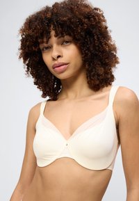 Frau mit lockigem Haar, die einen cremefarbenen BH trägt, leicht nach links blickend, vor einem einfarbigen hellgrauen Hintergrund.
