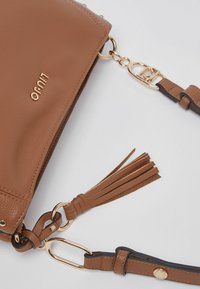 Sac bandoulière marron texturé avec ferrures dorées, accent pompon et sangle ajustable. Présente le nom de la marque embossé à l'avant.