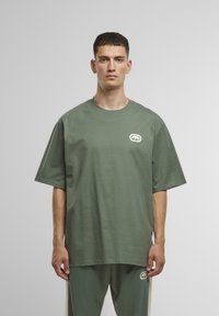 Overdimensioneret kortærmet t-shirt i olivengrøn bomuld, med et hvidt logo på venstre bryst og en glat, blød tekstur.