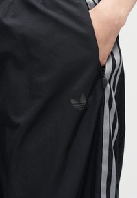Χέρι στην τσέπη μαύρου παντελονιού Adidas με γκρι λεπτομέρεια τριών ρίγων και φερμουάρ στο πλάι.