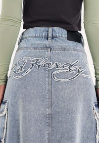 Lys blå denimnederdel med slidte detaljer, med "Ed Hardy" broderet på ryggen og en glat sort talje.