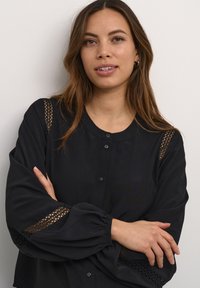 Melnas blūzes ar noapaļotu dekoltē, pogu priekšpusi, pūkainām piedurknēm un mežģīņu akcentiem uz pleciem un aprocēm. Gluda auduma tekstūra.
