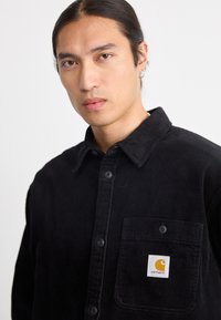 Un homme aux longs cheveux noirs attachés porte une chemise en velours côtelé noire à boutons avec un patch logo Carhartt sur la poche de la poitrine, sur fond blanc.