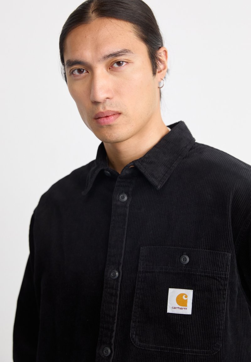 Man met lang, donker haar dat teruggebonden is, draagt een zwarte corduroy overhemdd met knopen en een Carhartt-logo patch op de borstzak tegen een witte achtergrond.