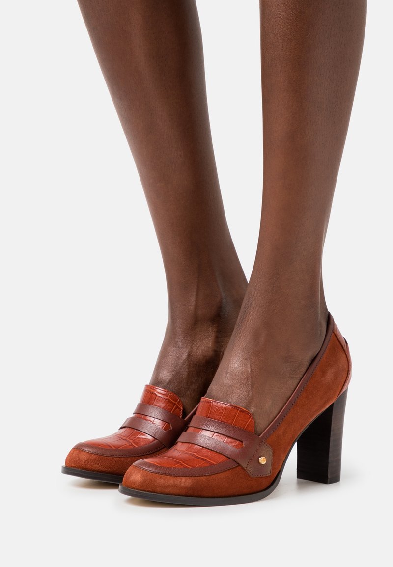 Mocassins à talons hauts en daim et cuir marron avec un motif texturé et un détail de lanière, portés aux pieds sur un fond uni.