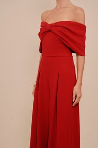 Ženska, oblečena v breznaramnico rdečo obleko z veliko zasukano pentljo na prsih, stoji ob enobarvni bež ozadju.