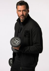 Sort zip-up jakke med præget "UNLMTD" branding, parret med en sort T-shirt og holder en sort håndvægt med hvide logo-accenter.