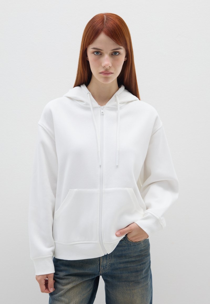 Sudadera blanca con cremallera y capucha, con bolsillos frontales, que presenta puños y dobladillo acanalados. Combinada con unos vaqueros de mezclilla azules. Textura suave, diseño sencillo.