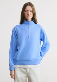 Νεαρό άτομο με μακριά μαλλιά που φοράει φούτερ με φερμουάρ έως το στήθος σε ανοιχτό μπλε χρώμα και λευκό παντελόνι, στέκεται μπροστά σε απλό λευκό φόντο.