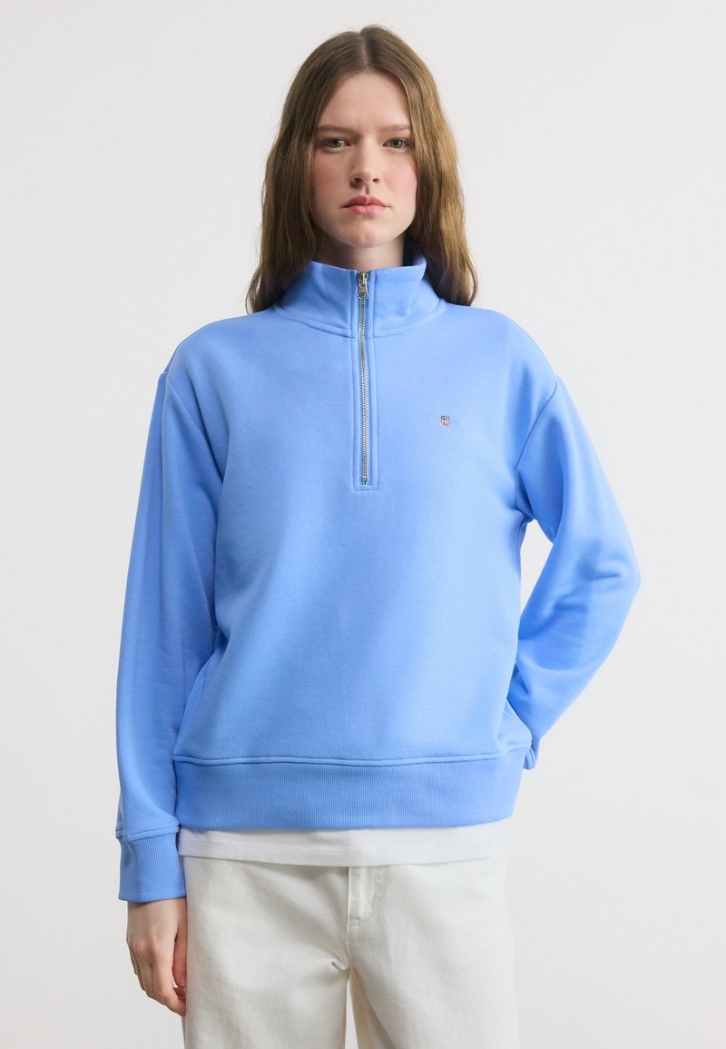 Νεαρό άτομο με μακριά μαλλιά που φοράει φούτερ με φερμουάρ έως το στήθος σε ανοιχτό μπλε χρώμα και λευκό παντελόνι, στέκεται μπροστά σε απλό λευκό φόντο.