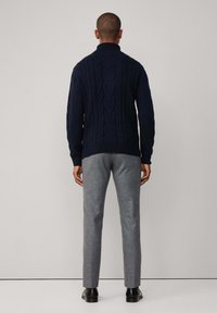 Muž oblečený v námořnickém modrém svetr s copánkovým vzorem, šedých upravených kalhotách a černých botách, stojící čelem k hladké světle šedé zdi.