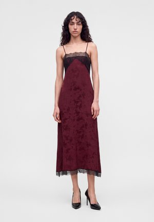Femme aux cheveux bouclés portant une robe midi bordeaux sans manches avec un bord en dentelle noire et des talons hauts noirs, debout sur un fond uni.