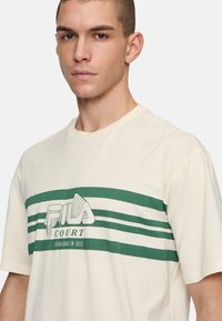 Valkoinen t-paita, joka on valmistettu puuvillasta, ja jossa on vihreä graafinen painatus, jossa on vaakasuoria raitoja ja keskellä teksti 'FILA COURT'.
