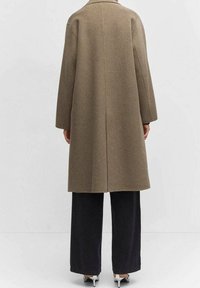 Cappotto di lana marrone con taglio dritto e maniche lunghe, caratterizzato da una cucitura centrale sul retro, abbinato a pantaloni neri a gamba larga.