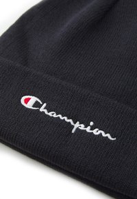 Gorro de malha preto com uma textura suave, apresentando um logótipo branco "Champion" e um pequeno emblema vermelho. Design dobrado.