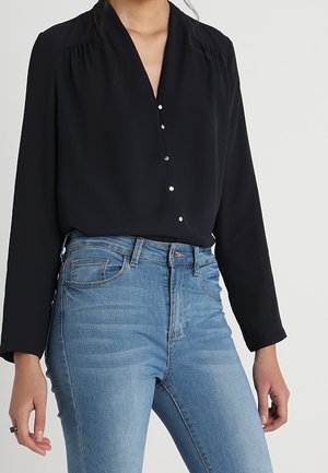 Blusa nera a maniche lunghe con scollatura a V e cinque piccoli bottoni sul davanti. Abbinata a jeans blu a vita alta, con una finitura sbiadita.