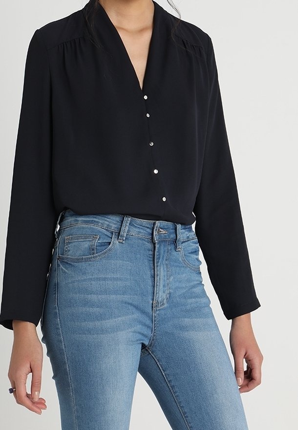 Blusa nera a maniche lunghe con scollatura a V e cinque piccoli bottoni sul davanti. Abbinata a jeans blu a vita alta, con una finitura sbiadita.