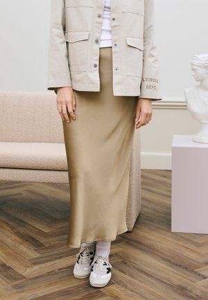 Persona con giacca beige, gonna color cuoio alla caviglia, calzini bianchi e scarpe da ginnastica bianche, in piedi su pavimento in legno accanto a un divano beige e a un busto bianco.