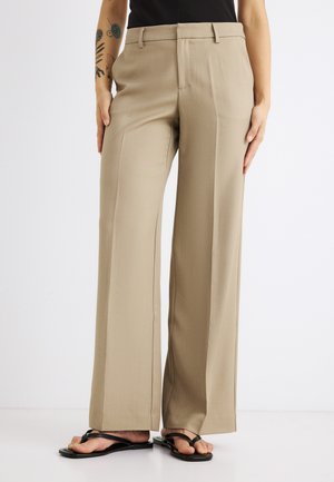 Smėlio spalvos platėjantys kelnės su diržo kilpomis, dėvimos su juodomis atvirų pirštų sandalais, parodytos nuo juosmens iki kojų prieš baltą foną.